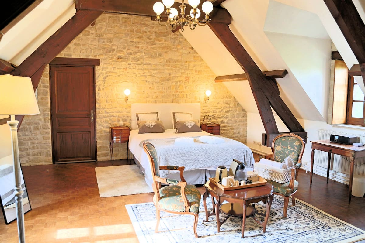 Een slaapkamer met een stenen muur, houten balken, een bed met kussens, een stoel, een tafel en een lamp.