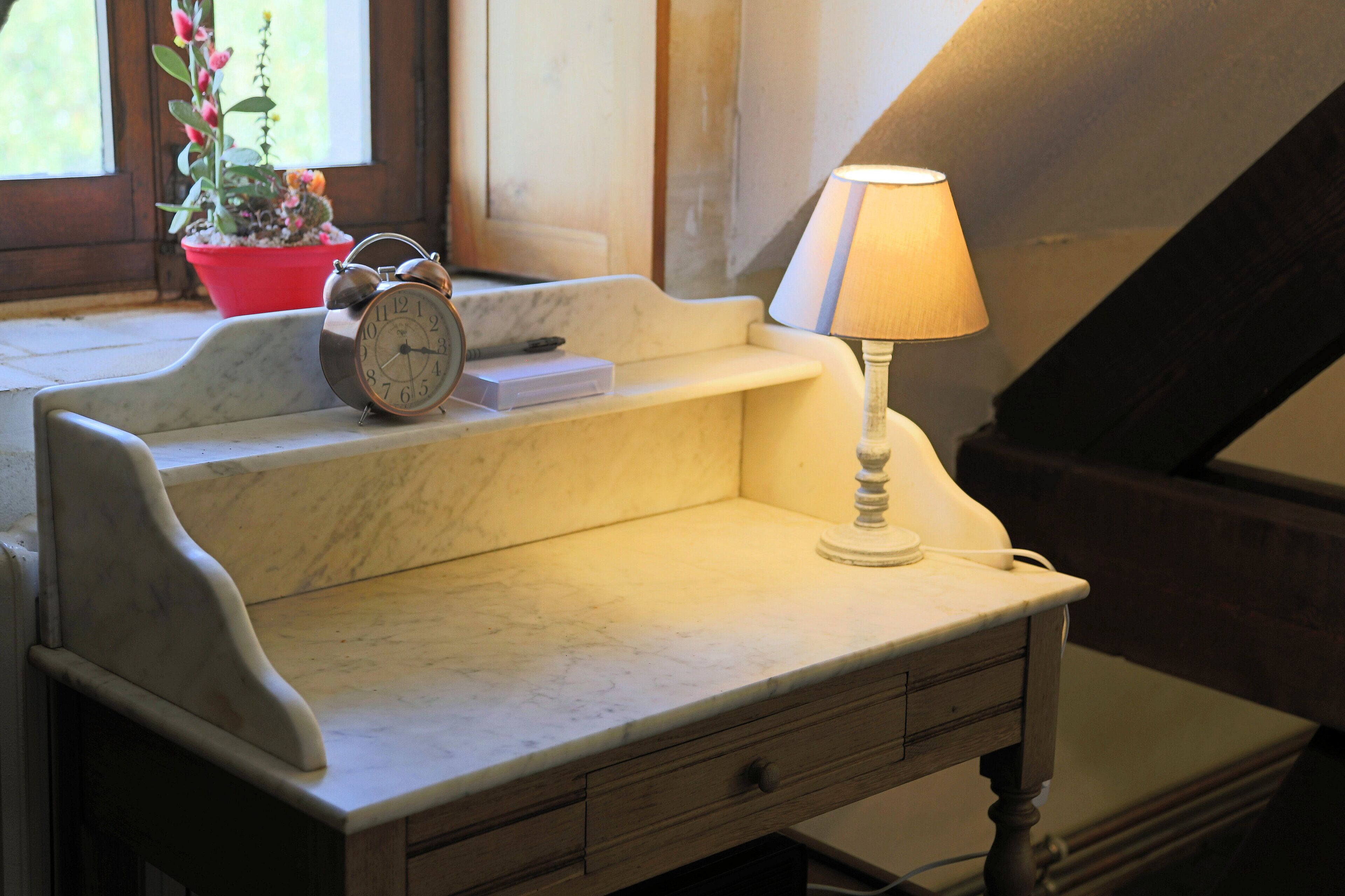 A marble desk with a lamp, a clock, and a potted plant by a window.