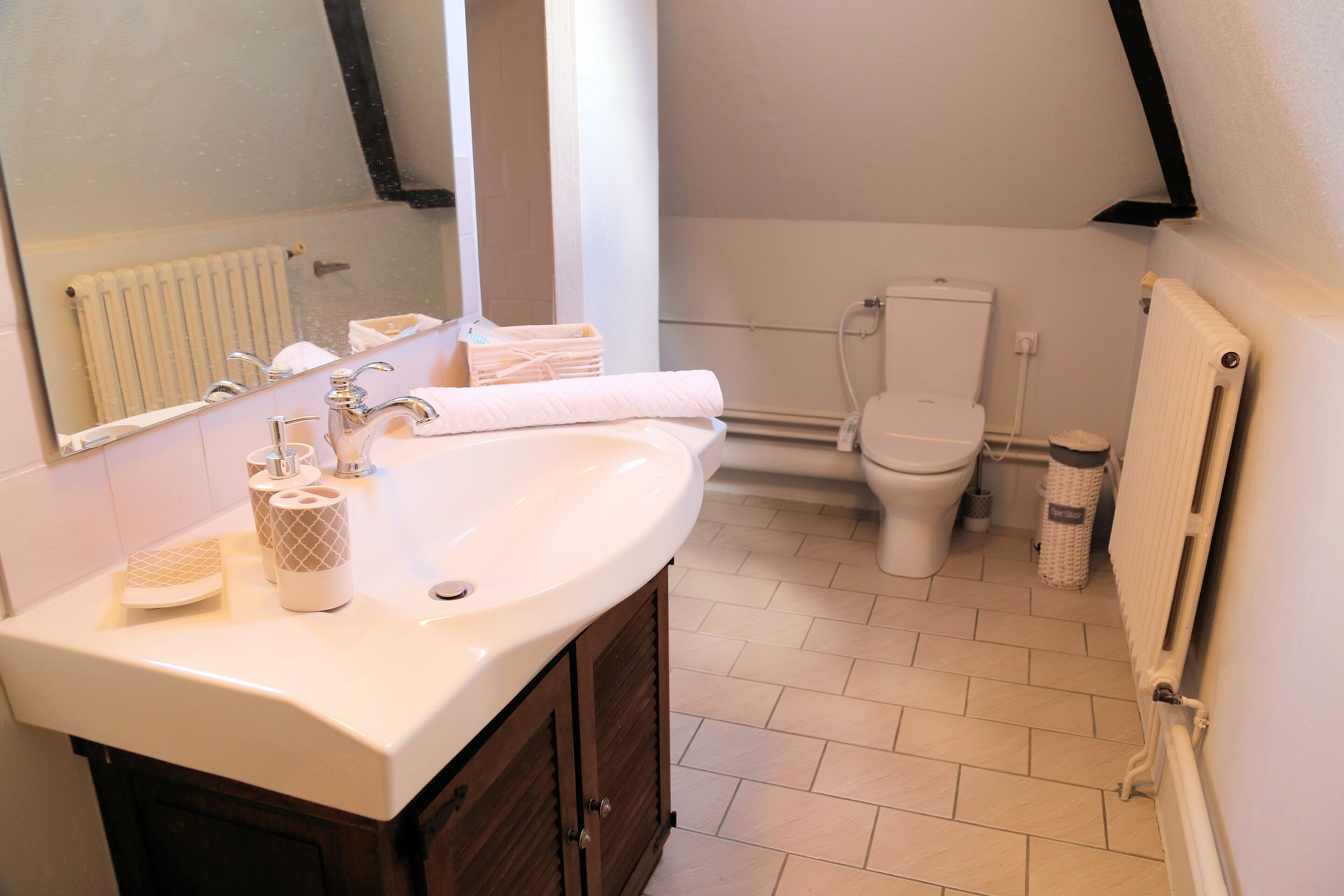 Une salle de bain avec un lavabo blanc, un miroir et des toilettes. On y trouve un porte-serviettes avec des serviettes et une corbeille à déchets.