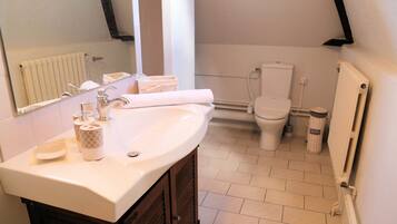 Une salle de bain avec un lavabo blanc, un miroir et des toilettes. On y trouve un porte-serviettes avec des serviettes et une corbeille à déchets.