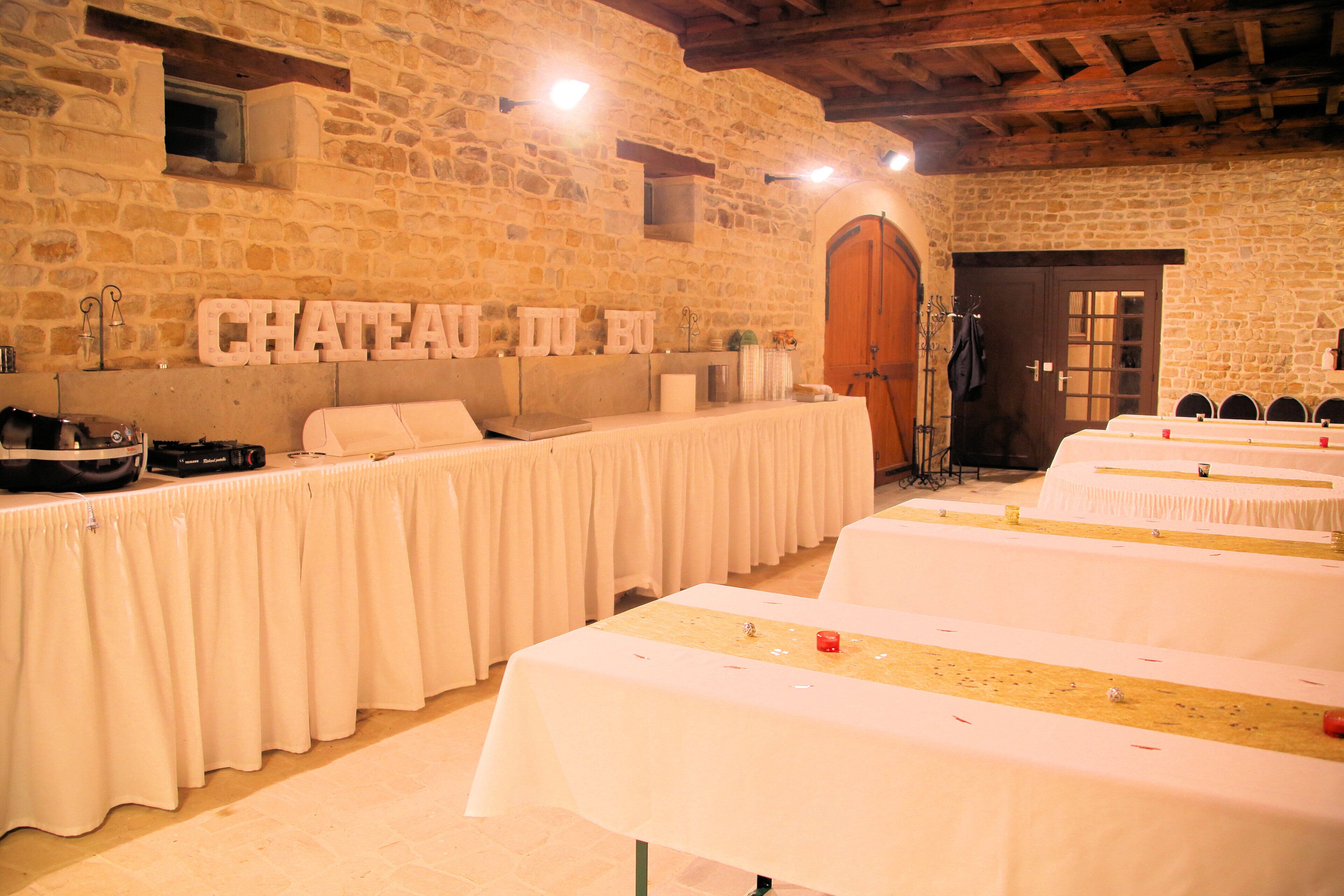 Un salón de banquetes con paredes de piedra y vigas de madera, mesas dispuestas para un evento formal, y un letrero que dice "CHATEAU DU BU".