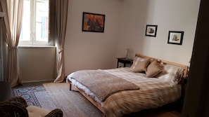 Ein Schlafzimmer mit einem Bett, einem Nachttisch, einer Lampe und einem Fenster mit Vorhängen.