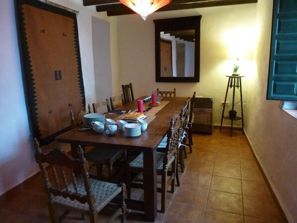 A dining area with a long wooden table set for six, surrounded by wooden chairs. A mirror hangs on the wall, and a small table with a lamp is visible in the corner.