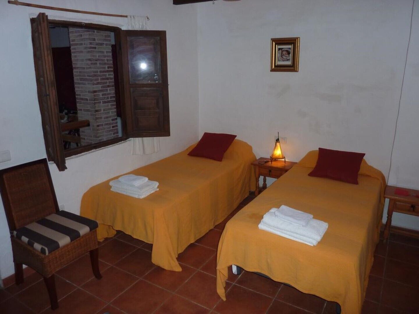 Una habitación con dos camas individuales, una silla de madera, una mesita y una ventana que da a un muro de ladrillo.