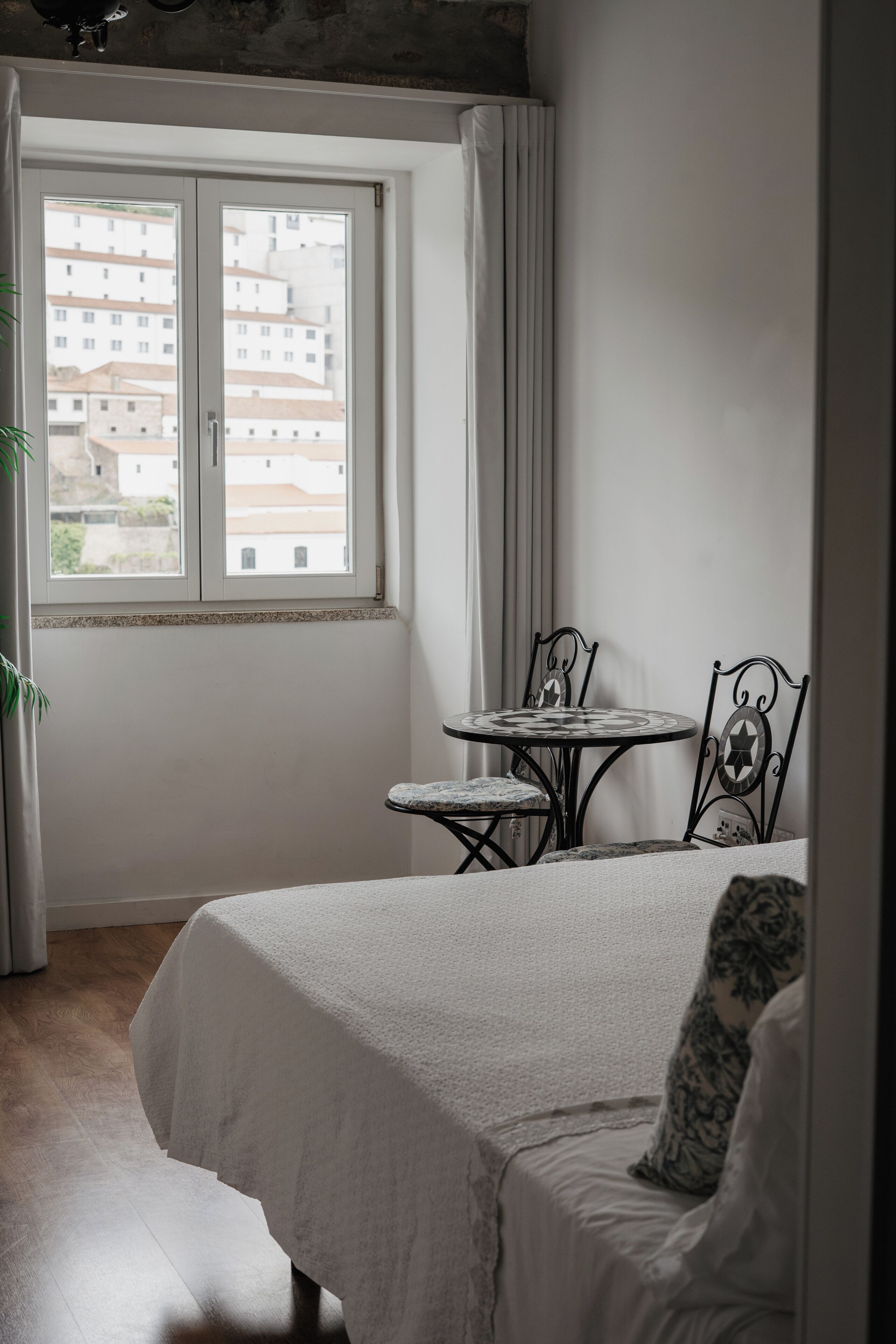 A bedroom with a bed, a small round table, two chairs, a window with curtains, and a view of buildings outside.