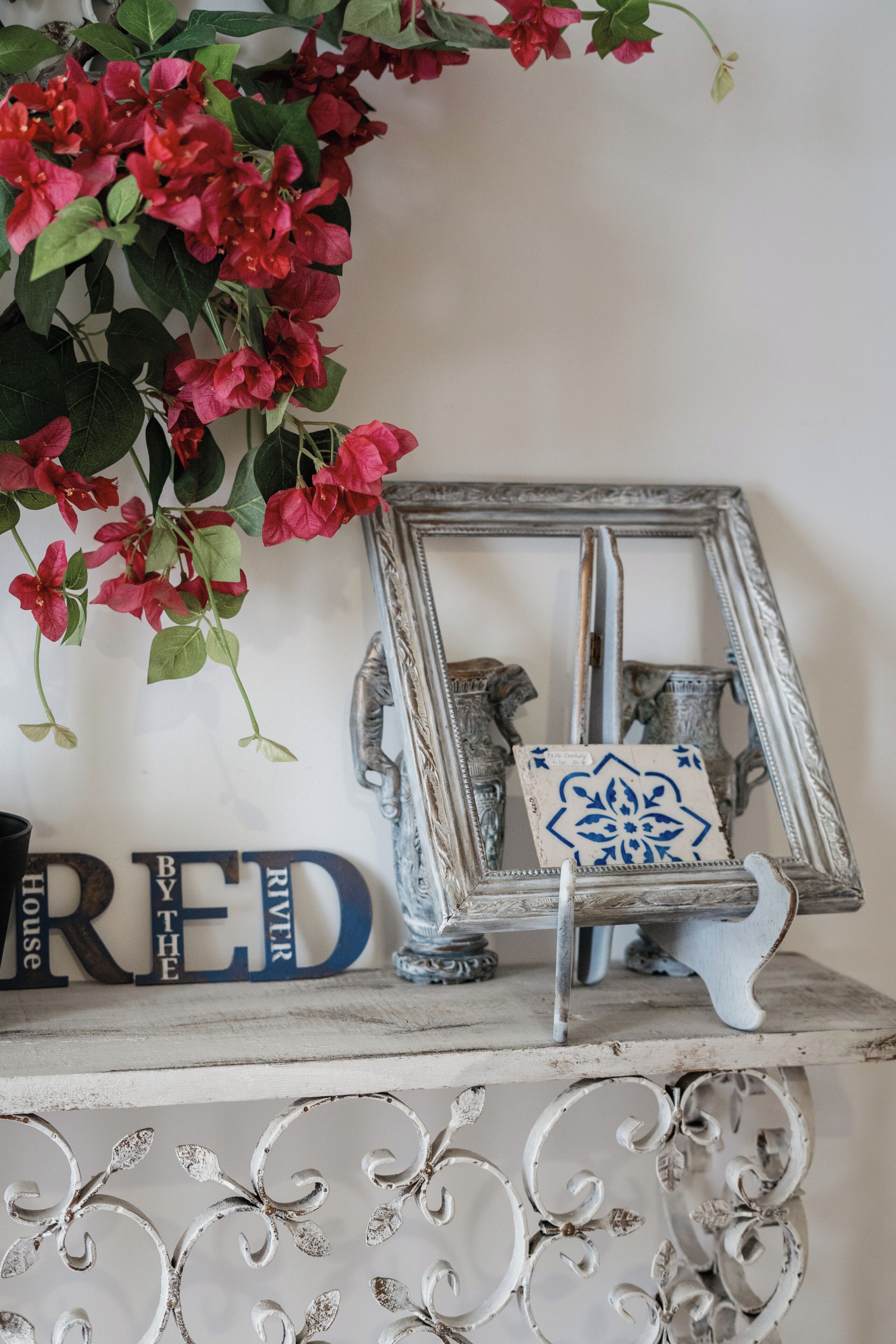 Una estantería decorativa con un espejo, un azulejo pequeño azul y blanco, y flores rojas.