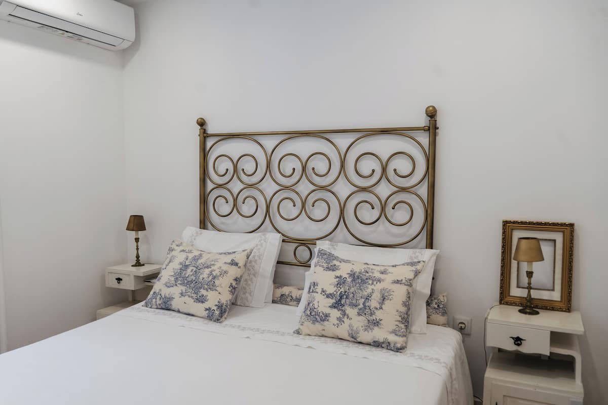 A bedroom with a bed, decorative headboard, bedside tables, and a wall-mounted air conditioner.
