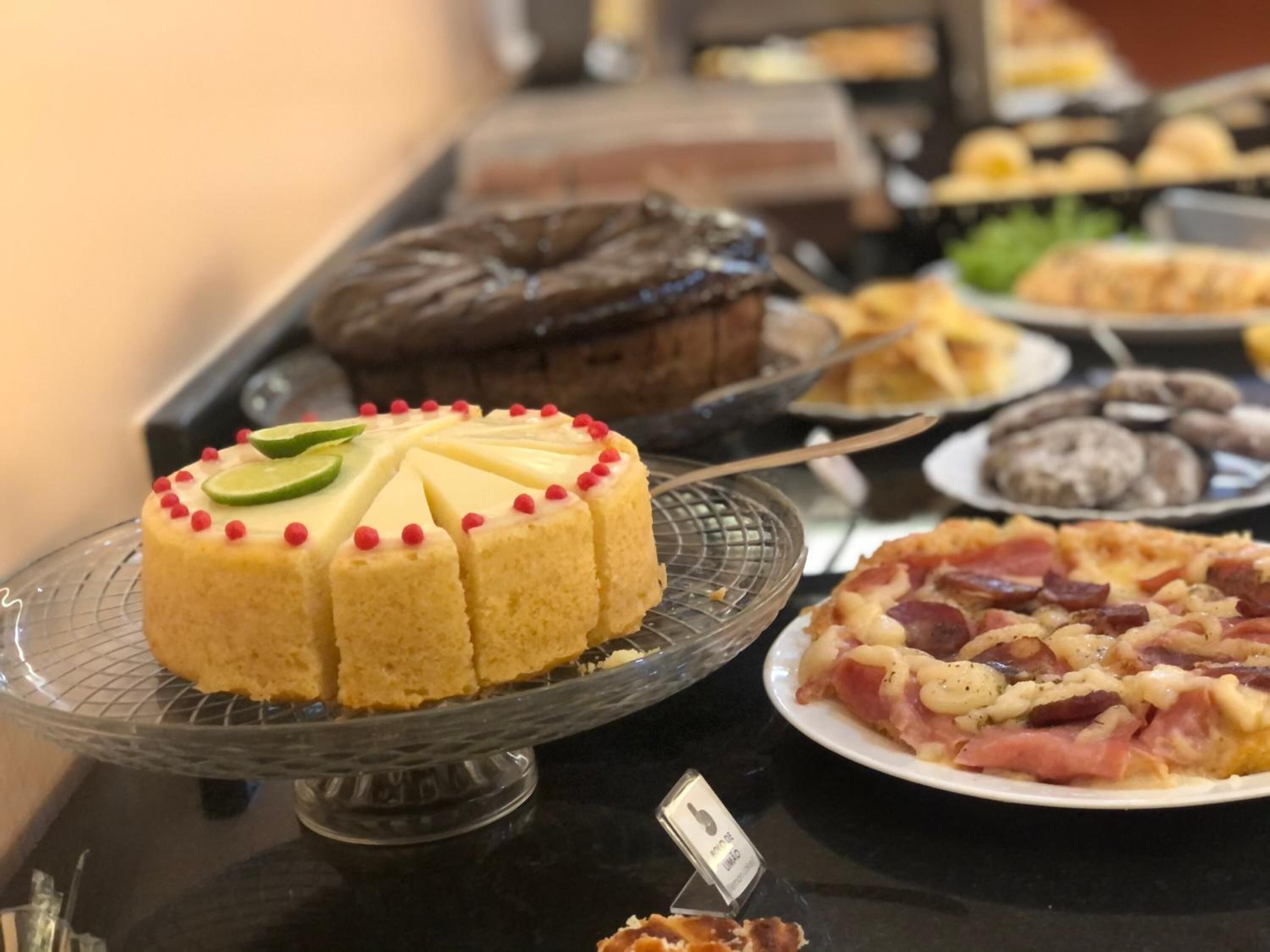 Una mesa de buffet con diversos platos, incluyendo un pastel con rodajas de lima y puntos rojos, un pastel de chocolate y una pizza de jamón y queso.