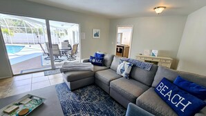 Ein Wohnzimmer mit grauem Sofa, Couchtisch und gemustertem Teppich. Durch die Schiebegläser ist ein Pool zu sehen.