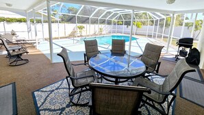 Ein überdachter Patio-Bereich mit Glastisch und -stühlen, einem Pool und Blick auf den Garten.