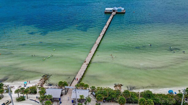 Luftaufnahme eines Piers, der sich in ein klares, blaues Meer erstreckt, während Menschen Wassersport betreiben.