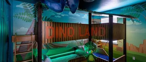 A children's playroom with a dinosaur theme, including a bed, a hanging dinosaur, and a mural.