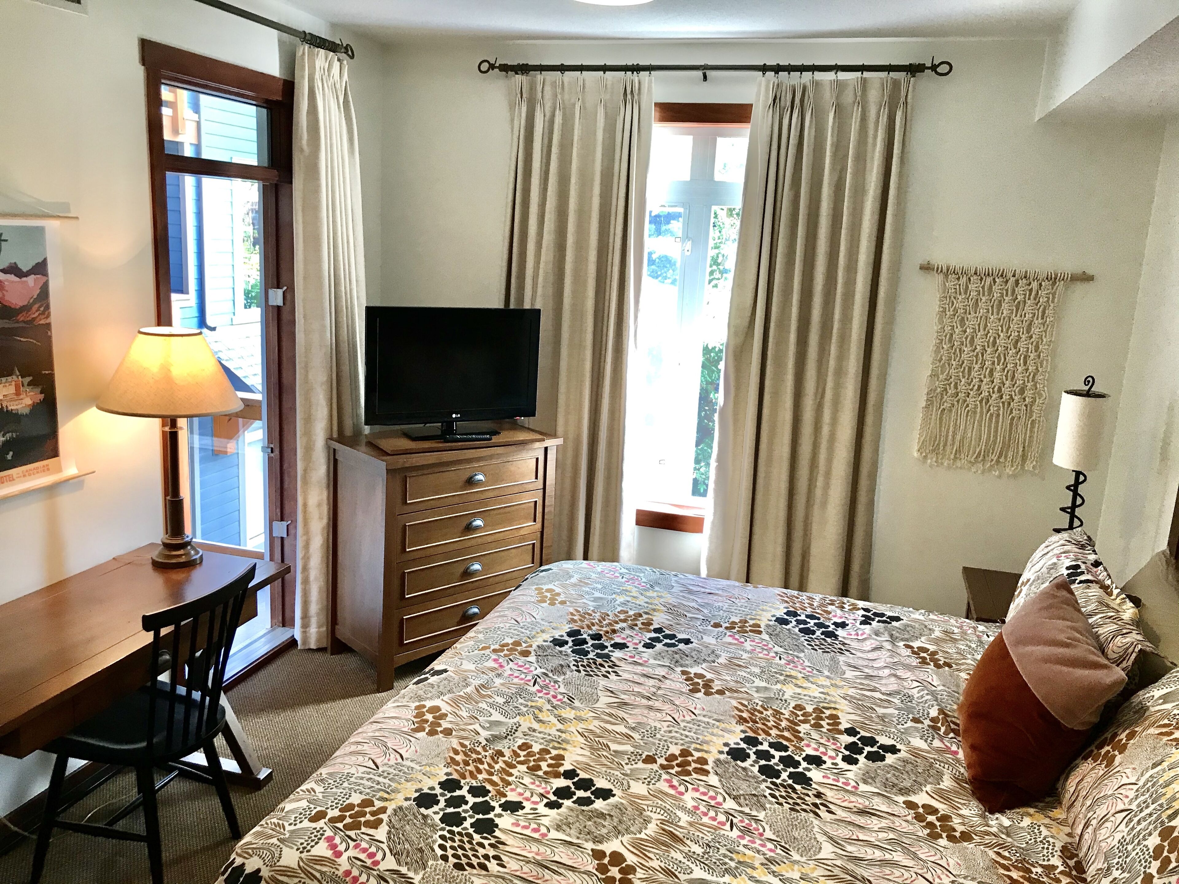 Quarto com cama, televisão sobre uma cômoda de madeira, escrivaninha com luminária e janela com cortinas.