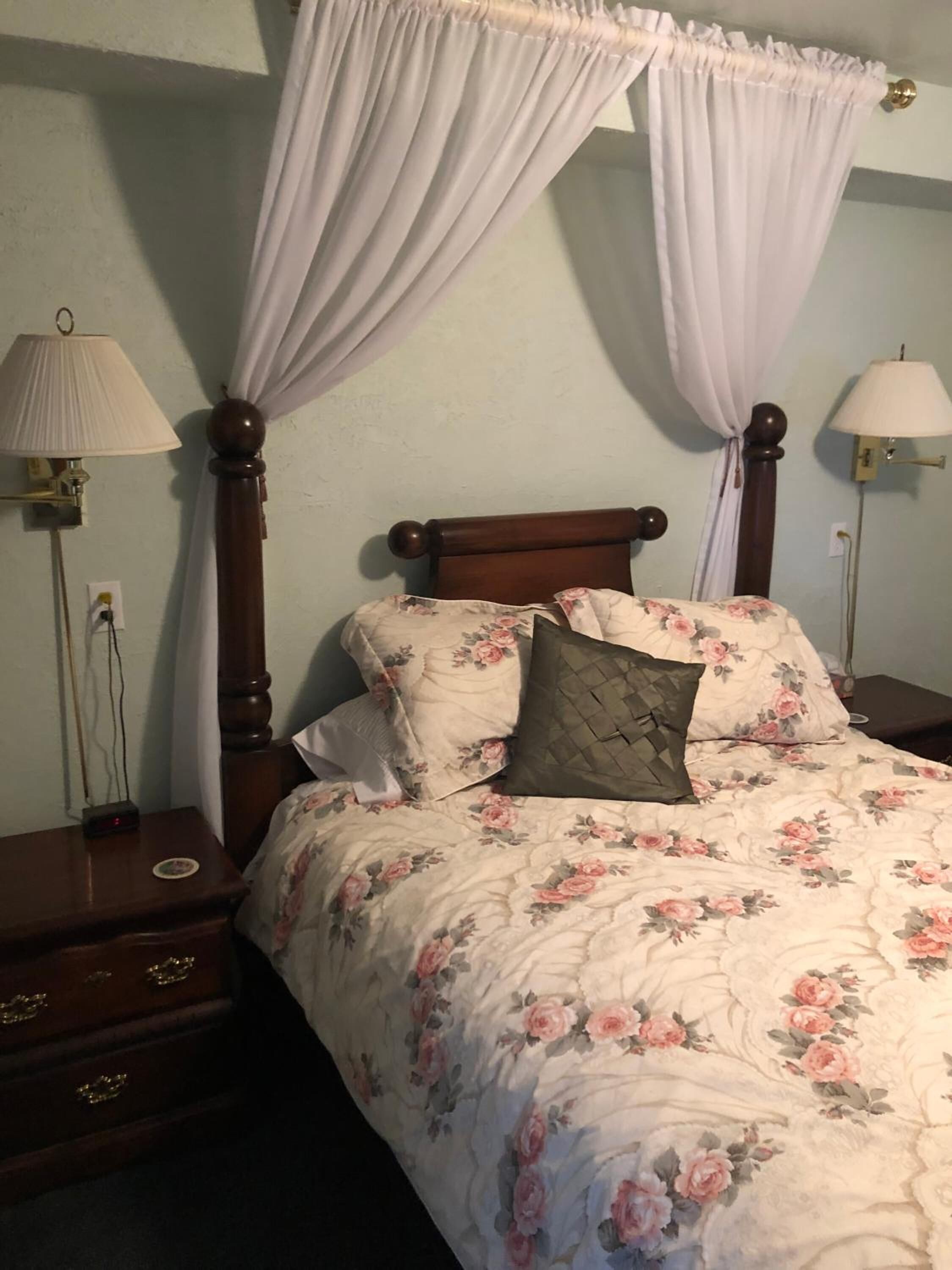 A bedroom with a four-poster bed, floral bedding, two bedside lamps, and a nightstand with drawers.