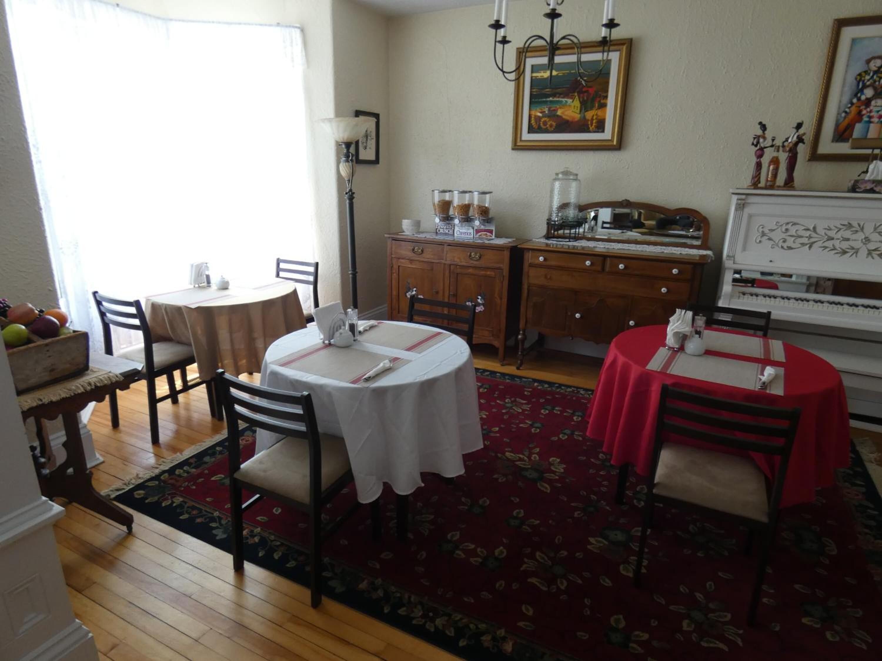 A dining area with two round tables, one with a white tablecloth and another with a red tablecloth, surrounded by wooden chairs. There is a wooden cabinet with glass jars and a framed painting on the wall.