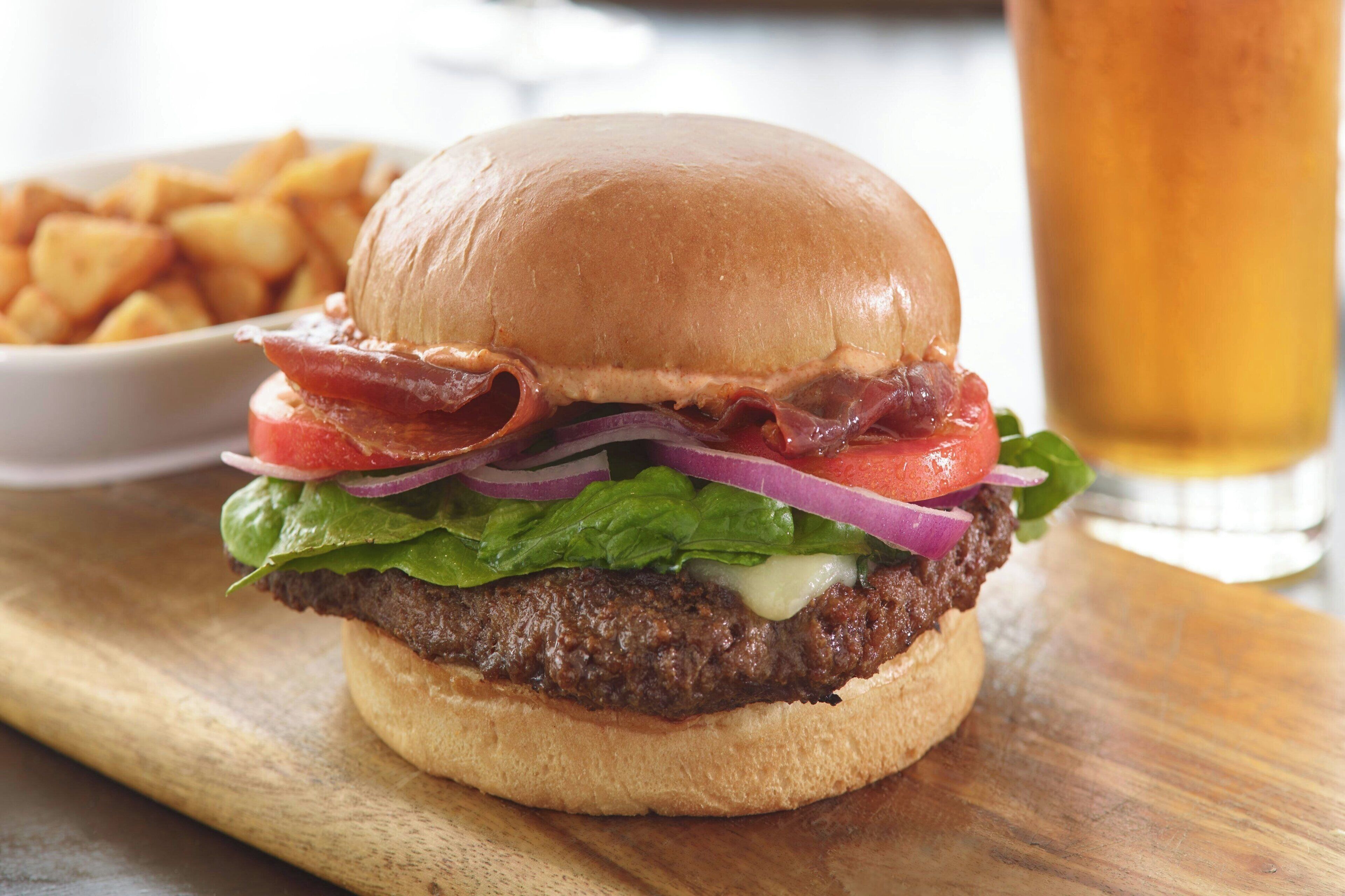 A burger with bacon, lettuce, tomato, and onion on a bun, served with fries and a glass of beer.
