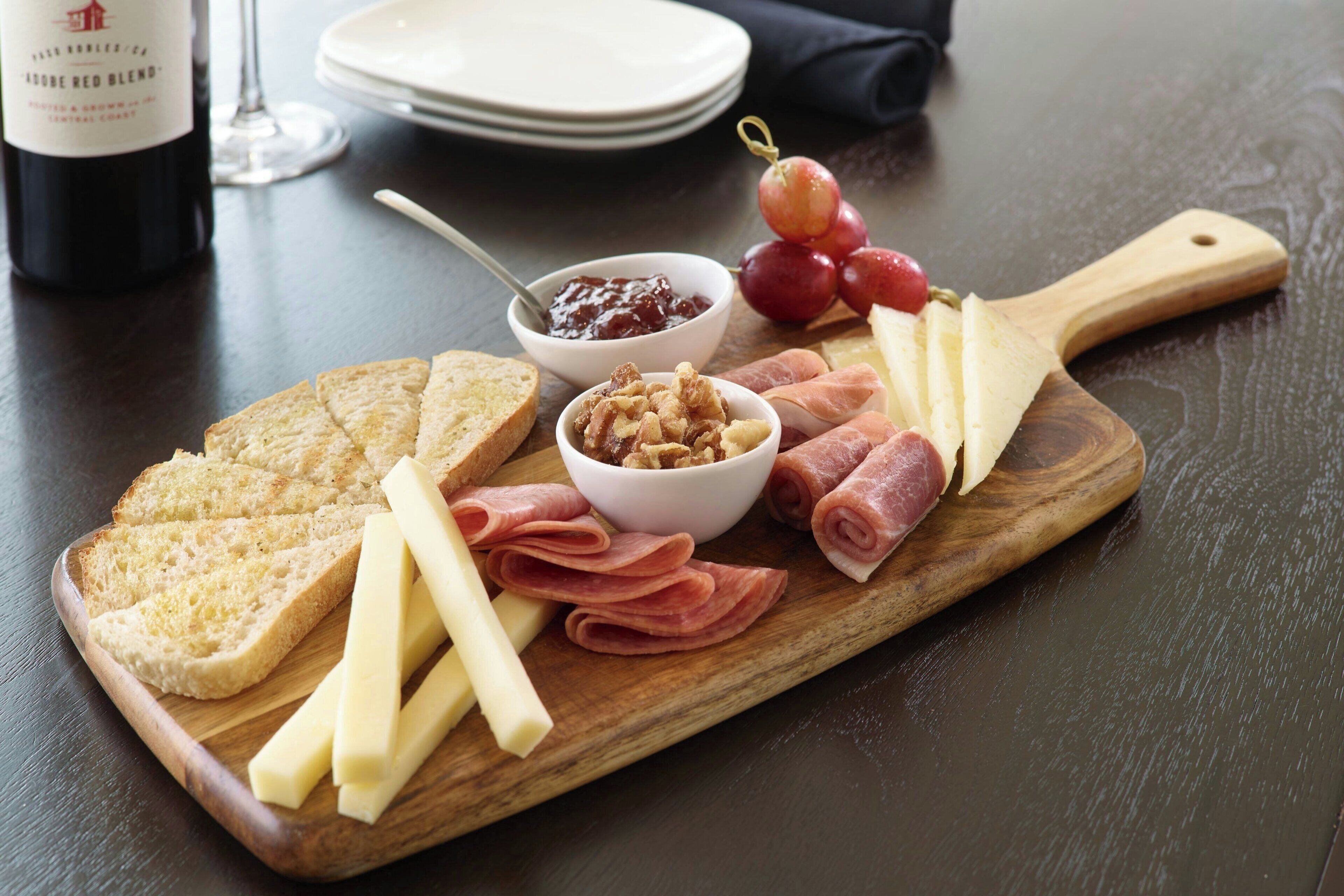 A wooden board with various cheeses, meats, and bread, accompanied by a bottle of wine and a bowl of jam.