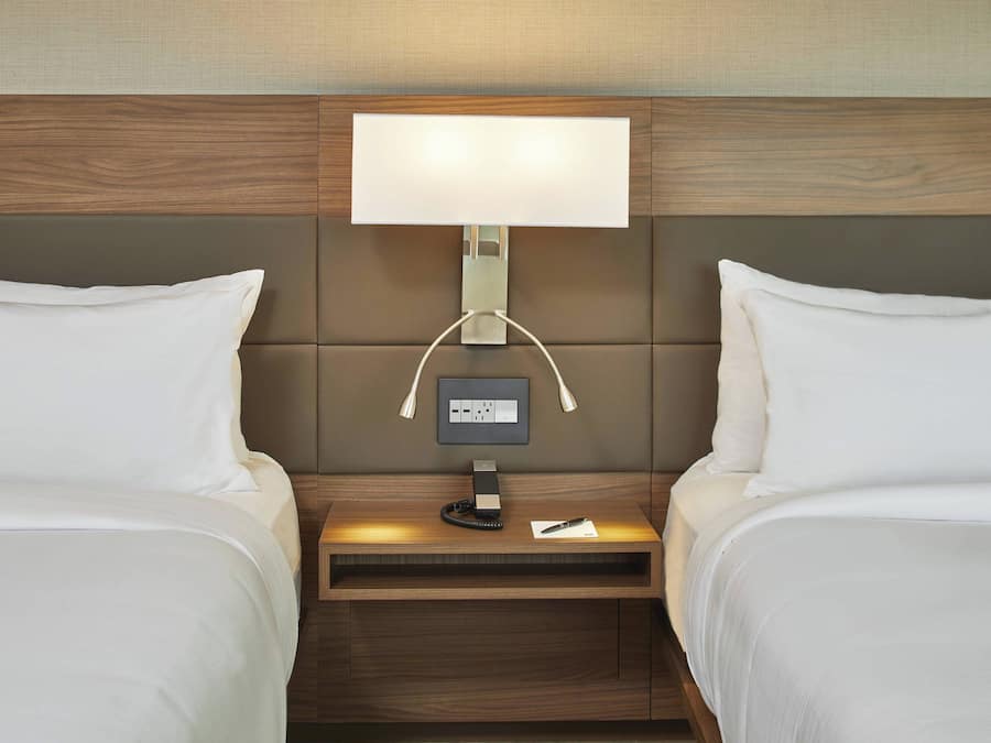 Habitación de hotel con dos camas, una mesita de noche con lámpara y un enchufe en la pared.