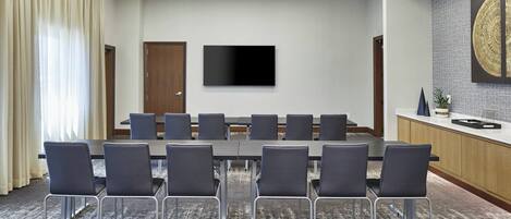 A conference room with a long table, chairs, a flat-screen TV, and a kitchen area with a decorative wall piece.