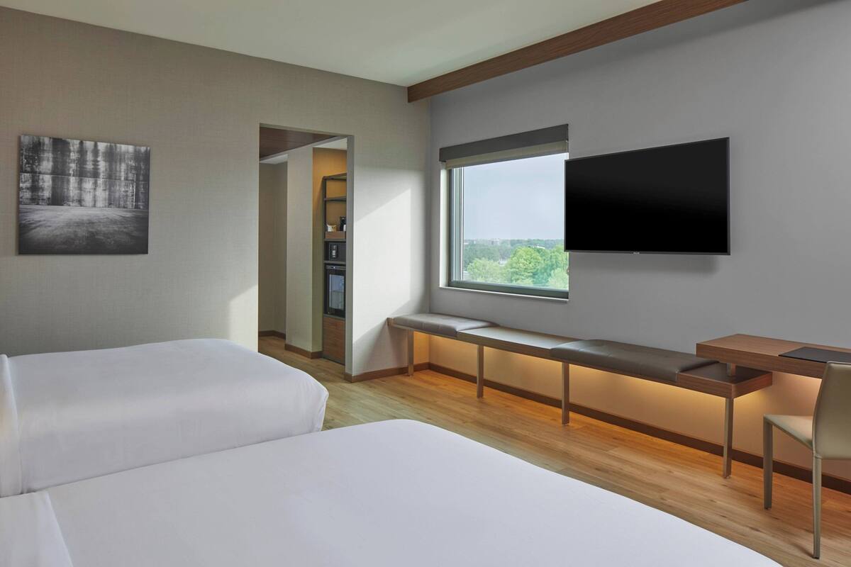 Een moderne hotelkamer met twee bedden, een bureau met stoel, een aan de muur bevestigde televisie en een raam met uitzicht op het landschap.