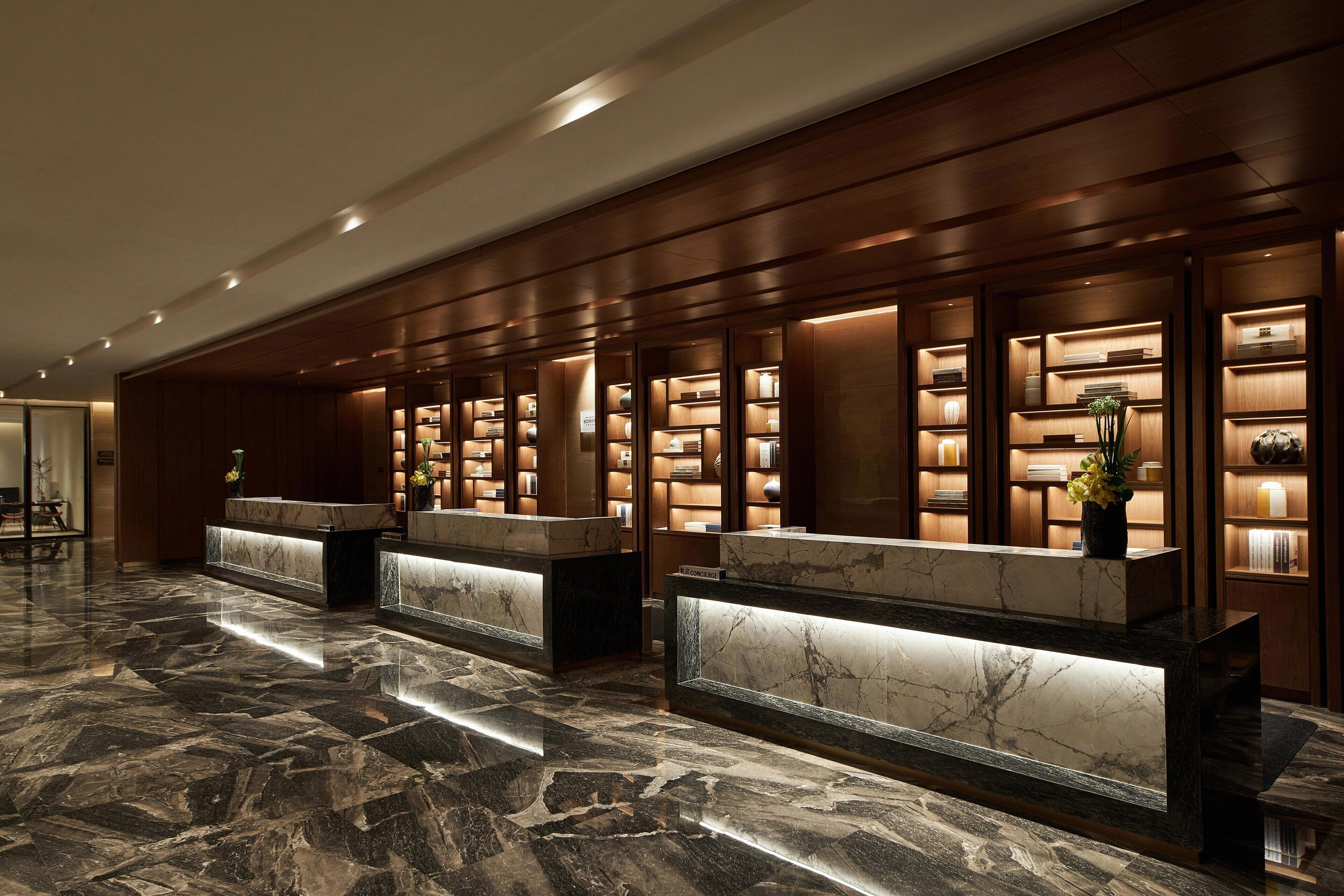 Zona de recepción de hotel con mostradores de mármol, paneles de madera y estanterías iluminadas que exhiben libros y objetos decorativos.