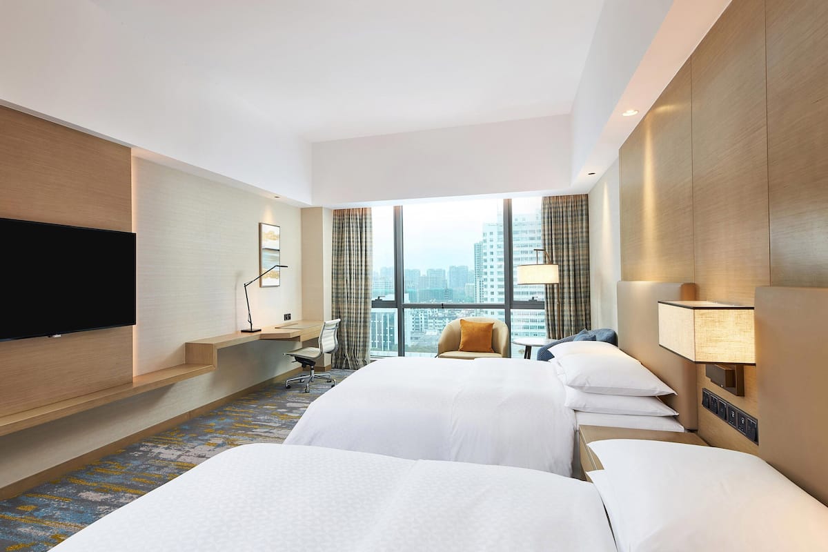 A hotel room with two beds, a desk, a chair, a TV, and a large window with a city view.