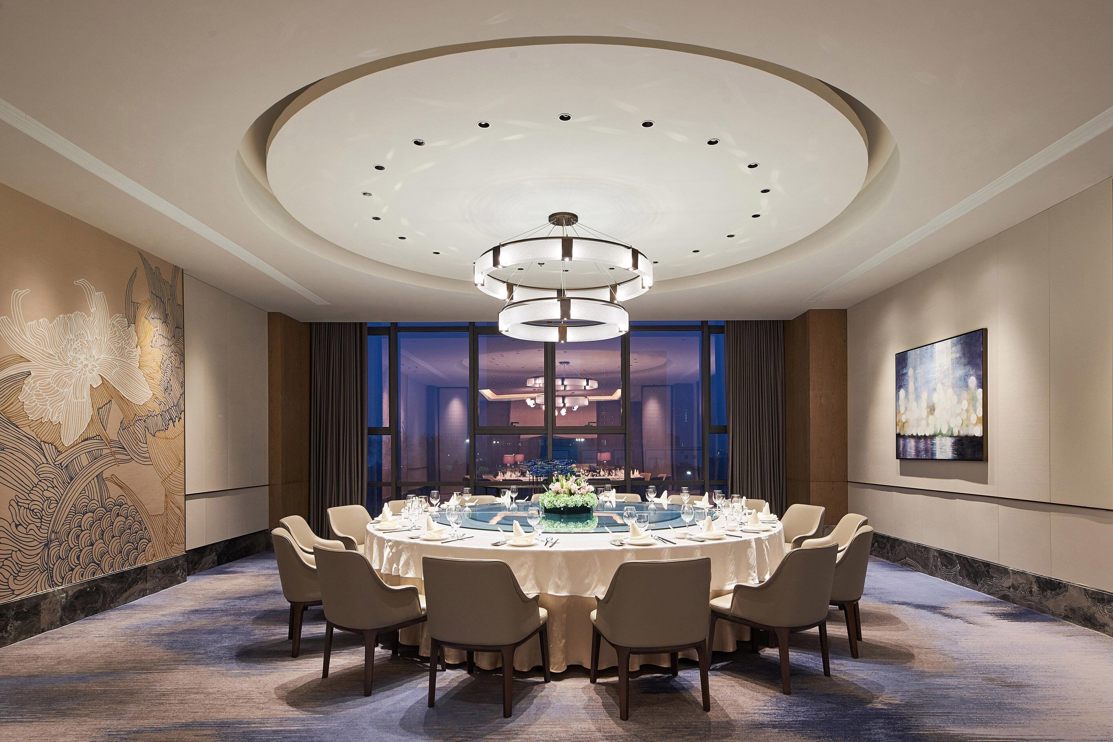 A round dining table set for six with a chandelier above, surrounded by chairs, in a room with large windows and a mural on the wall.