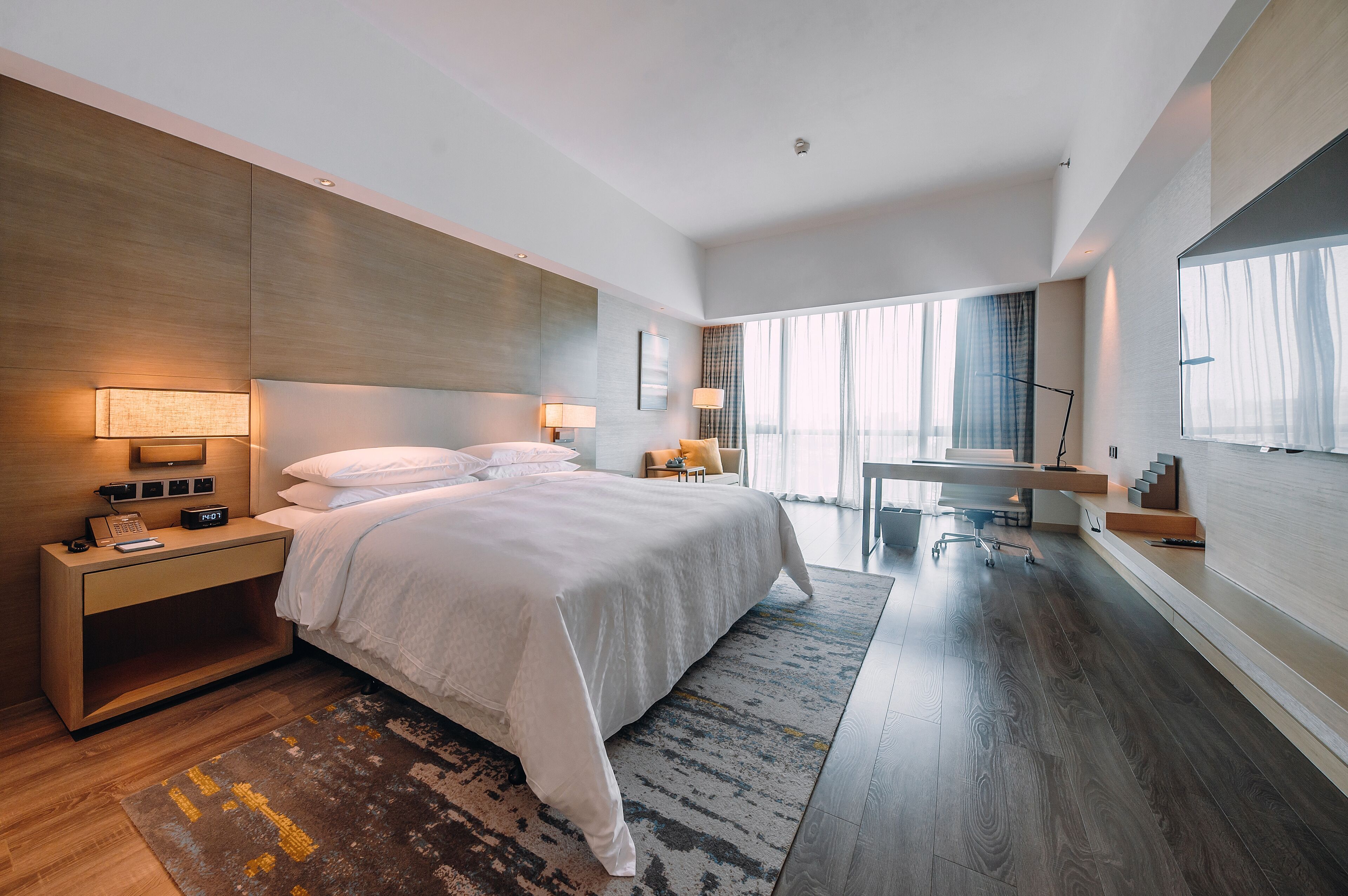 Una habitación de hotel moderna con una cama grande, lámparas de noche, un escritorio y vistas de la ciudad a través de amplias ventanas.
