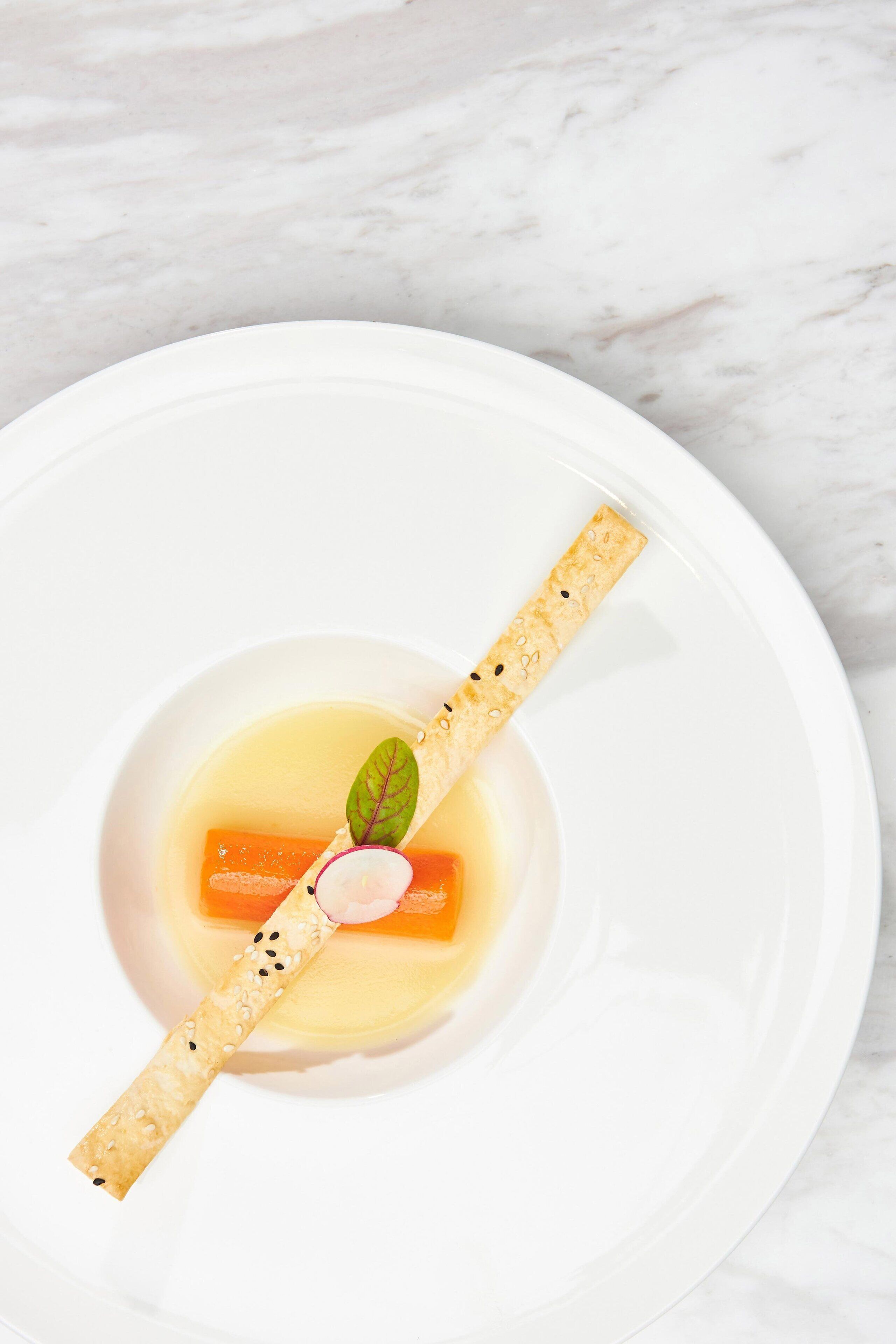 A white plate with a clear liquid, a carrot, and a cracker with seeds.
