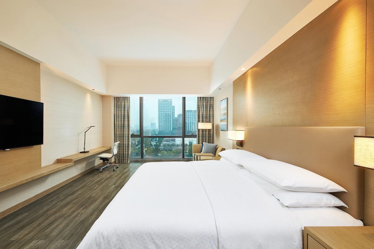 A modern hotel room with a large bed, a desk with a chair, a flat-screen TV, and a city view through large windows.