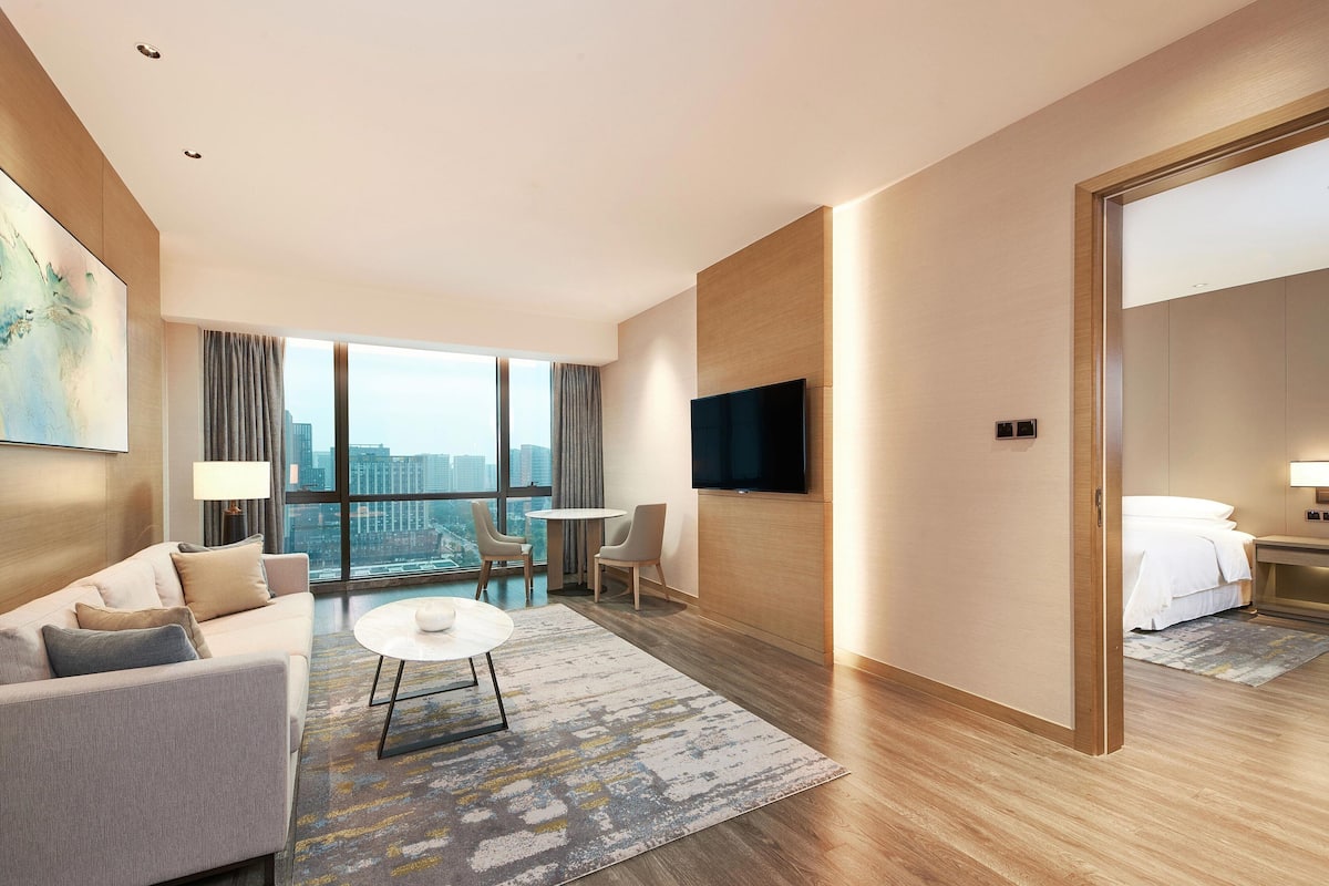 A modern hotel room with a sofa, chairs, a TV, and a large window with a city view.
