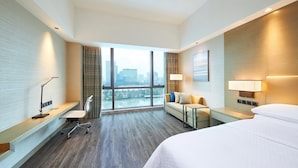 Habitación de hotel moderna con una cama grande, un escritorio con silla, un sofá y vistas de la ciudad a través de amplios ventanales.