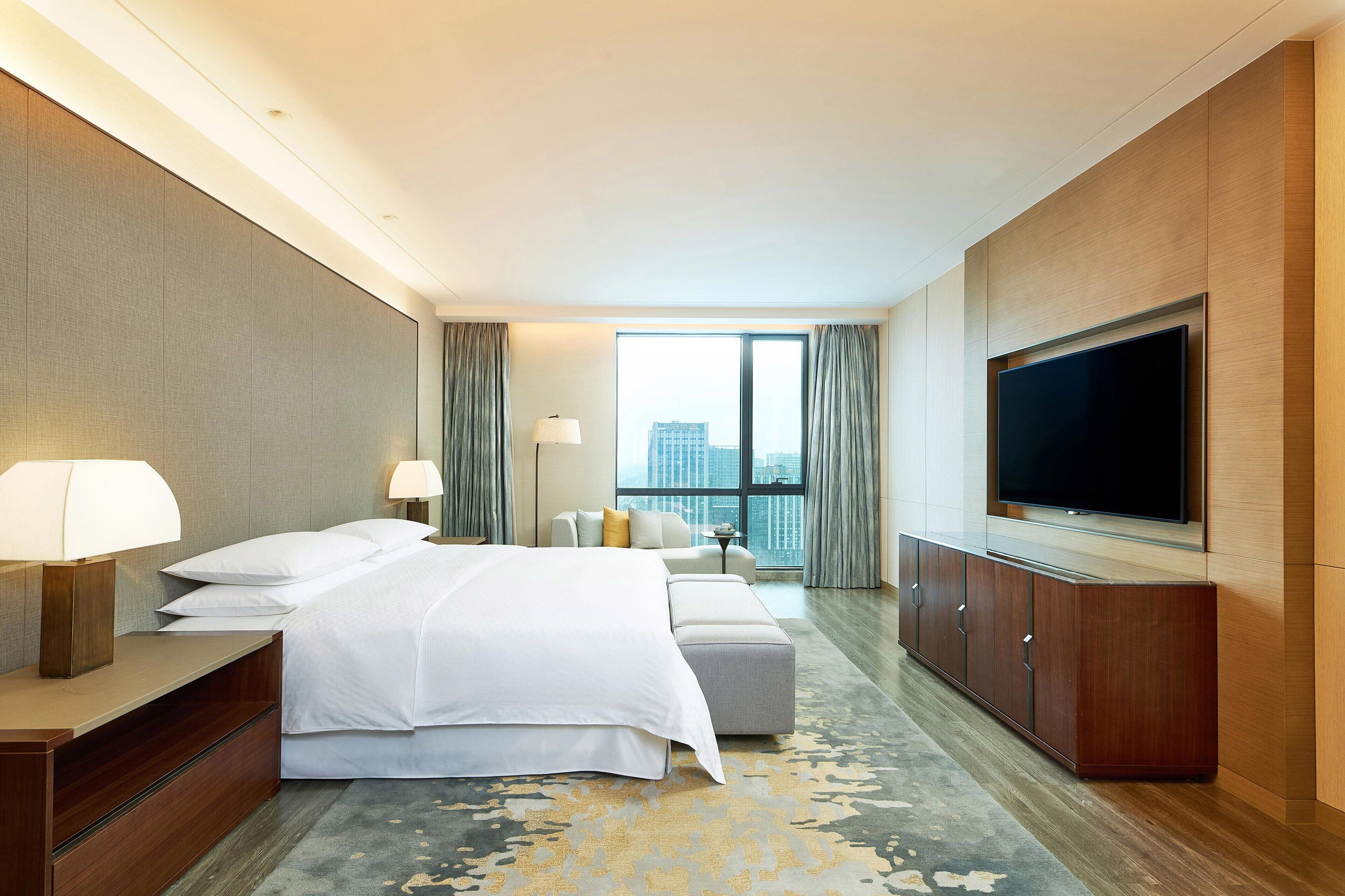 A modern hotel room with a large bed, a flat-screen TV, a sofa, and a view of the city.