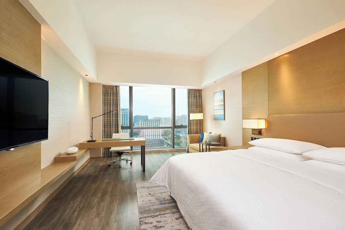 A hotel room with a large bed, a desk, a flat-screen TV, and a city view.