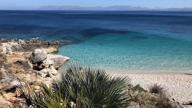 A coastal landscape with clear turquoise waters, rocky terrain, and sparse vegetation.