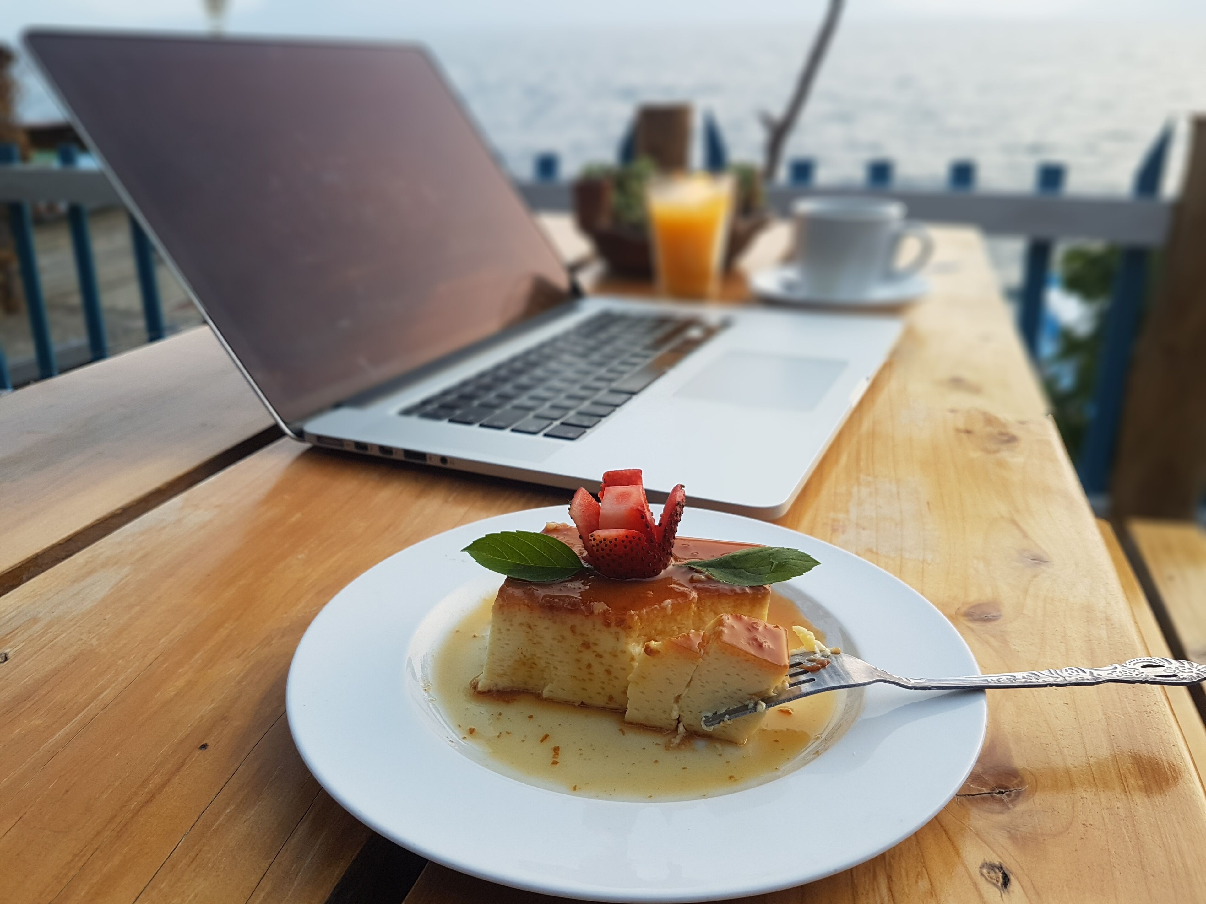 Sobremesa em um prato branco com um garfo, um laptop, uma xícara e um copo de suco de laranja em uma mesa de madeira perto de um corrimão.