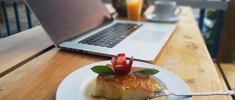 Sobremesa em um prato branco com um garfo, um laptop, uma xícara e um copo de suco de laranja em uma mesa de madeira perto de um corrimão.
