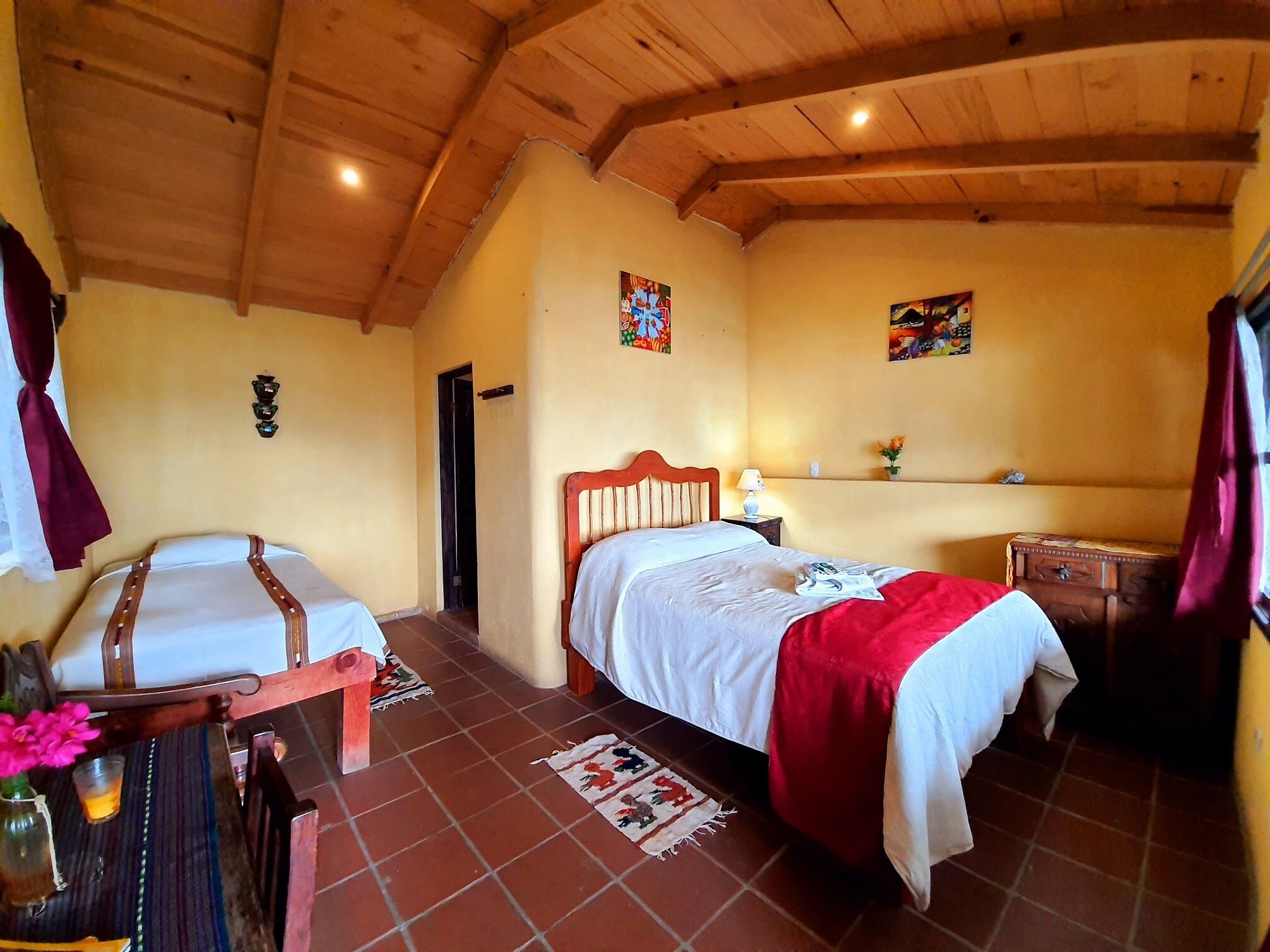 Une pièce chaleureuse avec un plafond en bois, un lit recouvert d’une couette rouge et blanche, une petite table ornée d’un vase et d’une tasse, et une chaise avec un coussin à rayures.