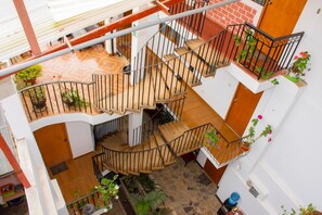 Un interior de varios niveles con escaleras de madera, balcones con plantas en macetas y piso de baldosas.