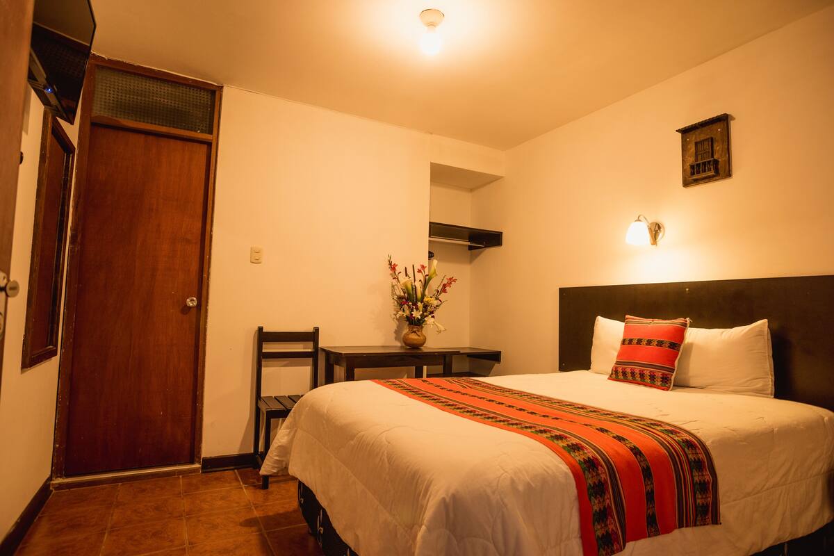 Une chambre d’hôtel avec un lit, un bureau agrémenté d’un vase de fleurs, une porte en bois et un luminaire fixé au mur.
