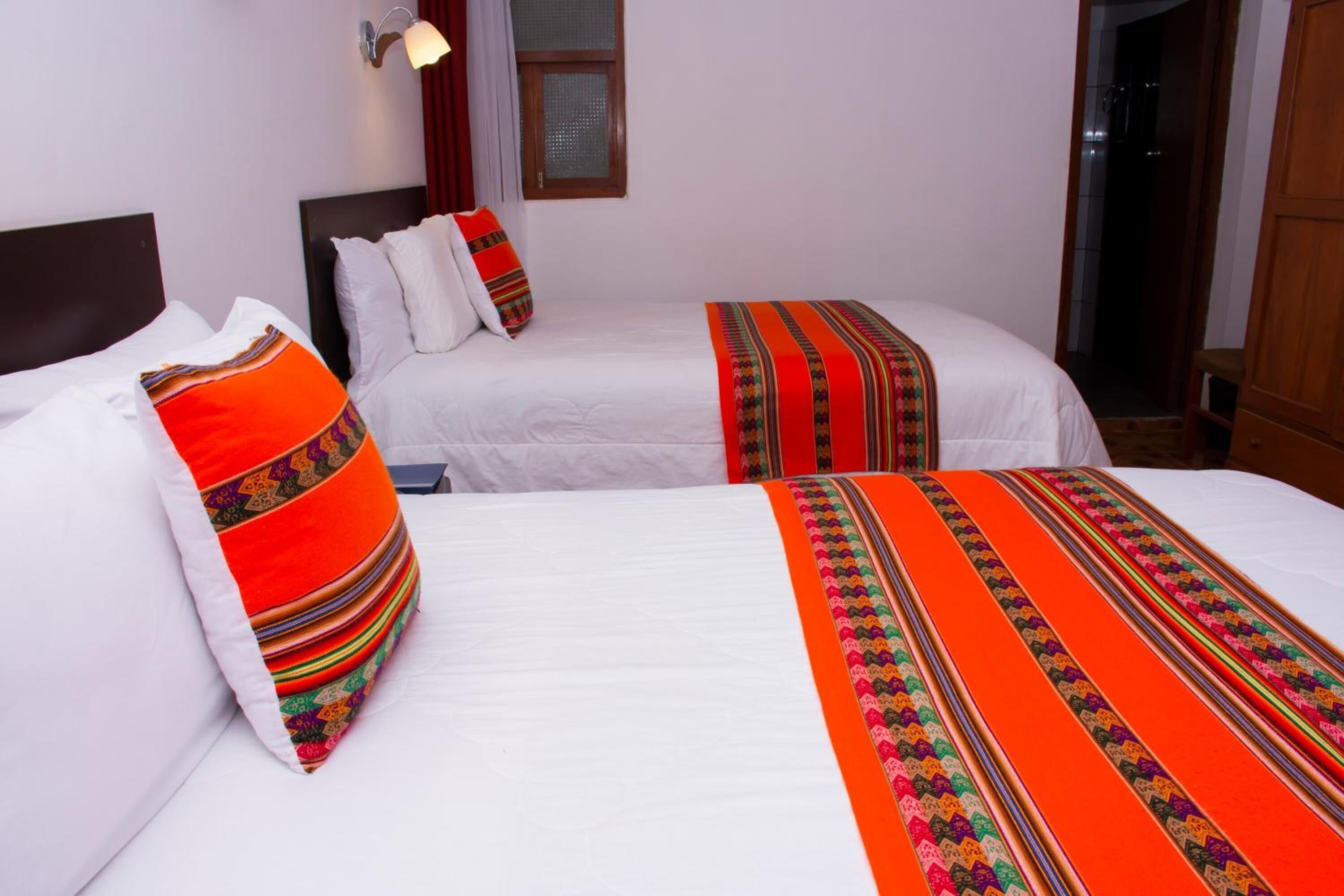 Habitació d'hotel amb dos llits, cadascun amb un estovat blanc i almoinades de colors. Hi ha una finestra amb cortines i un lluminària fixat a la paret.