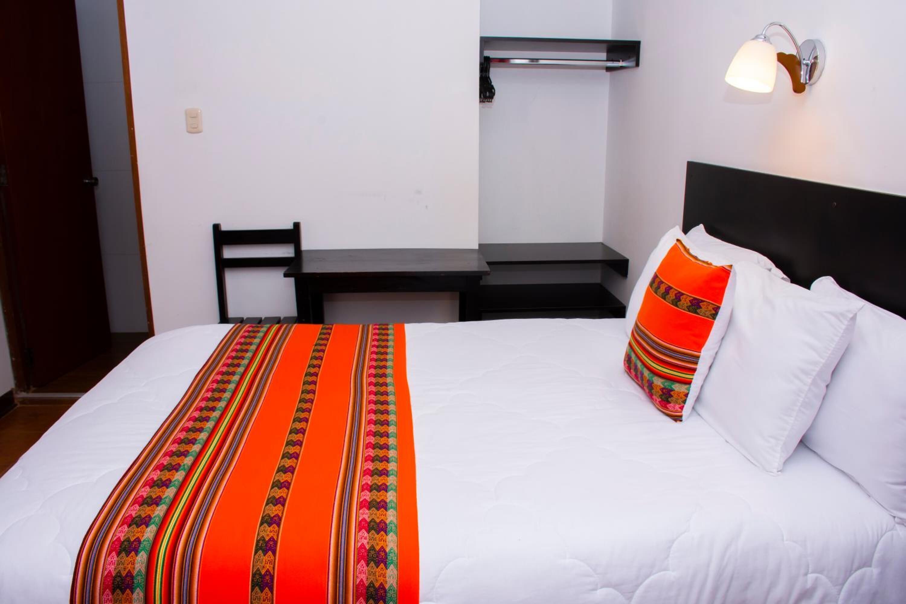 A neatly made bed with a white comforter and an orange and green striped blanket. There is a black headboard, a white pillow, and a red pillow with a striped pattern. A small table with a black surface is positioned under a wall-mounted light fixture.