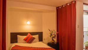A neatly made bed with a white comforter and a red and black patterned throw, a nightstand with a vase of flowers, and a red curtain partially drawn.