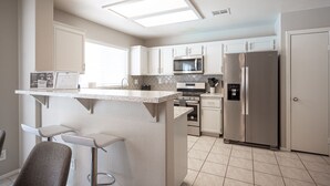 Una cocina moderna con armarios blancos, un refrigerador de acero inoxidable, un microondas y un horno empotrado. Cuenta con una barra con taburetes.