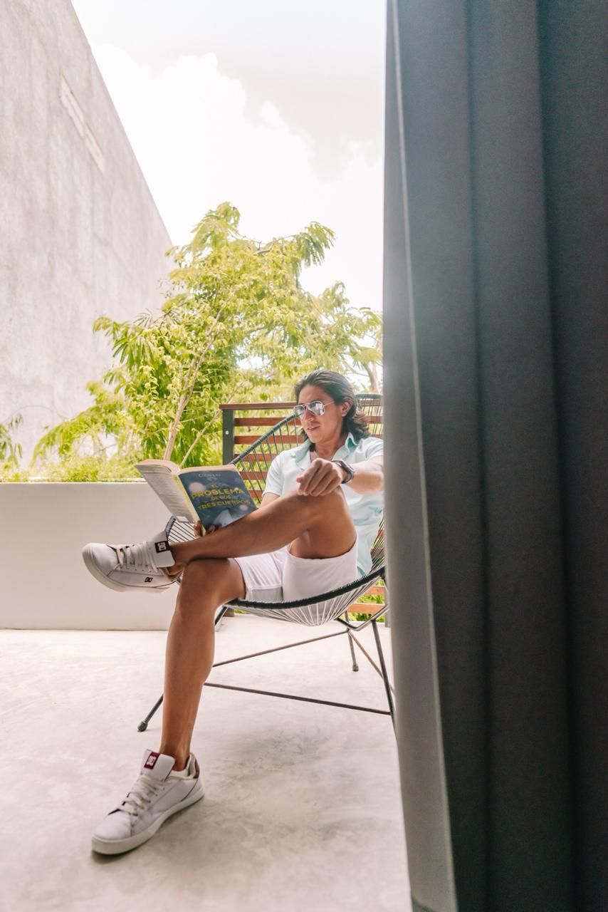 Une personne assise sur une chaise, en train de lire un livre, avec un mur en béton et de la verdure en arrière-plan.