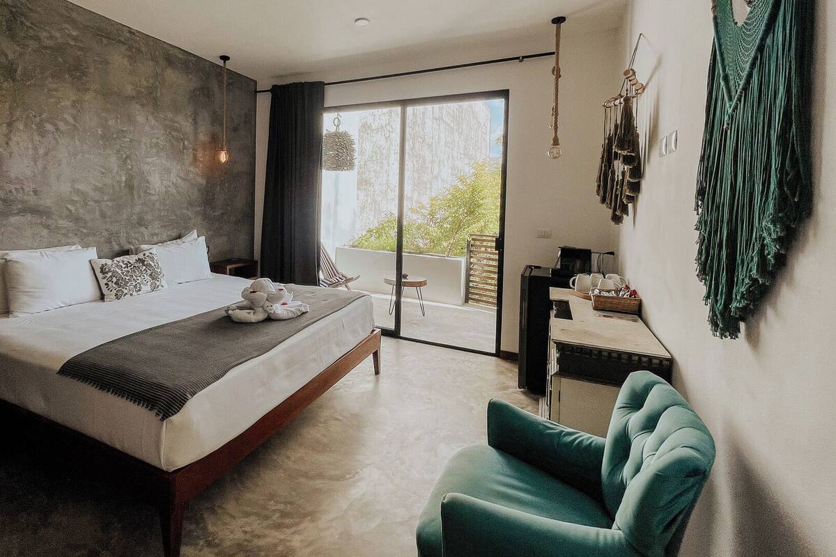 A modern bedroom with a large bed, a green armchair, and a view of greenery outside.
