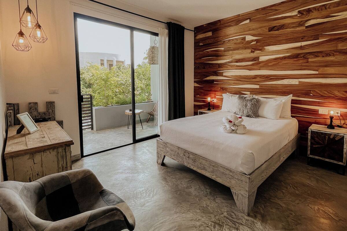 A bedroom with a large bed, a wooden accent wall, a view of greenery outside, and a small table with a lamp.