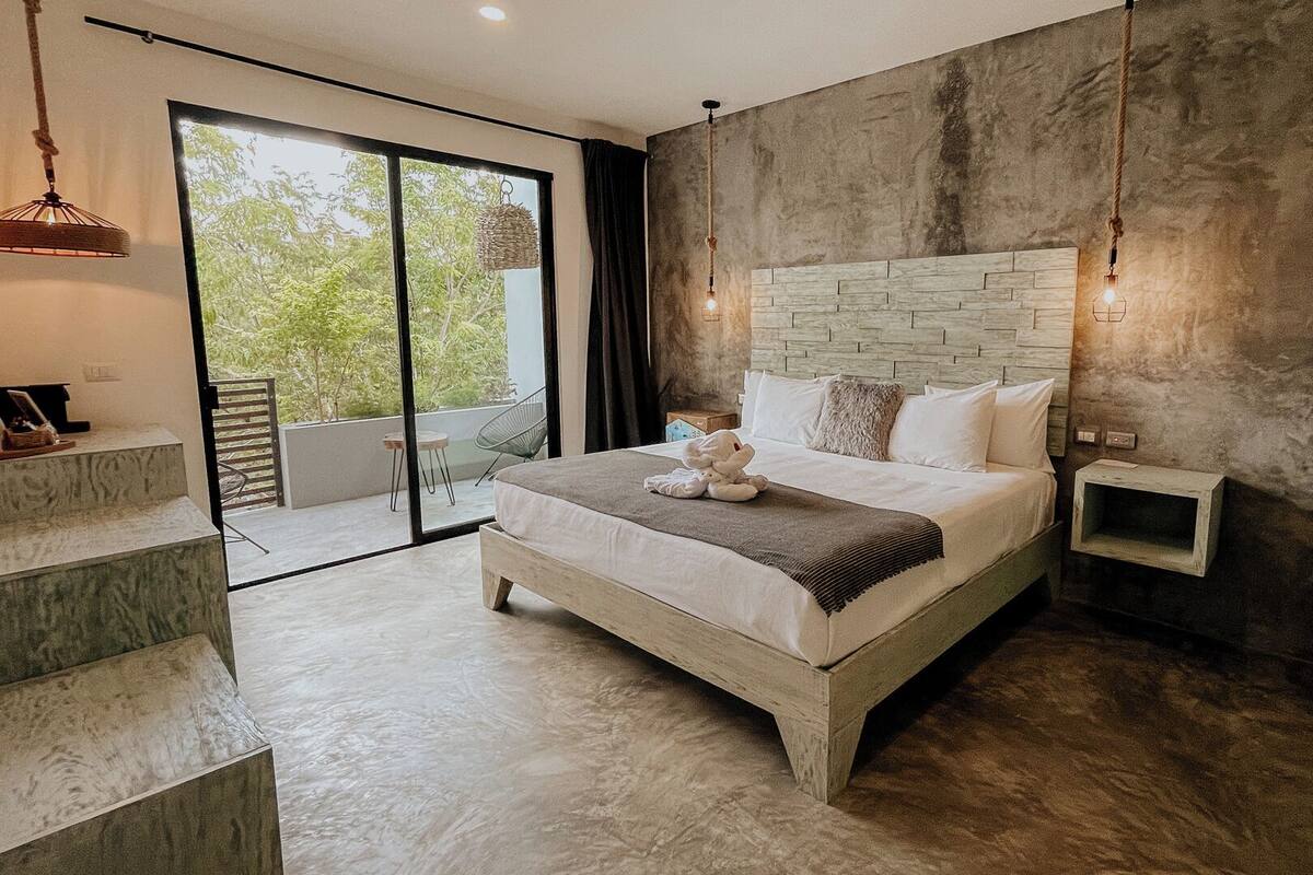 A modern bedroom with a stone headboard, a large bed, a bedside table, and a view of greenery through a sliding door.