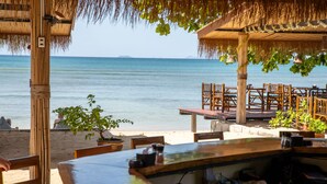 Ein Restaurant direkt am Strand mit Holztischen und -stühlen, einem Reetdach und Blick auf das Meer.