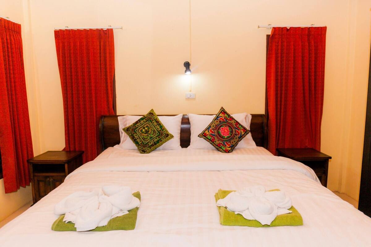 Une chambre d’hôtel avec un lit, des rideaux rouges, deux coussins décoratifs, une table de chevet et une lampe fixée au mur.